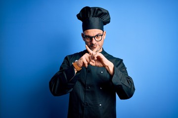Young handsome chef man wearing cooker uniform and hat over isolated blue background Rejection expression crossing fingers doing negative sign