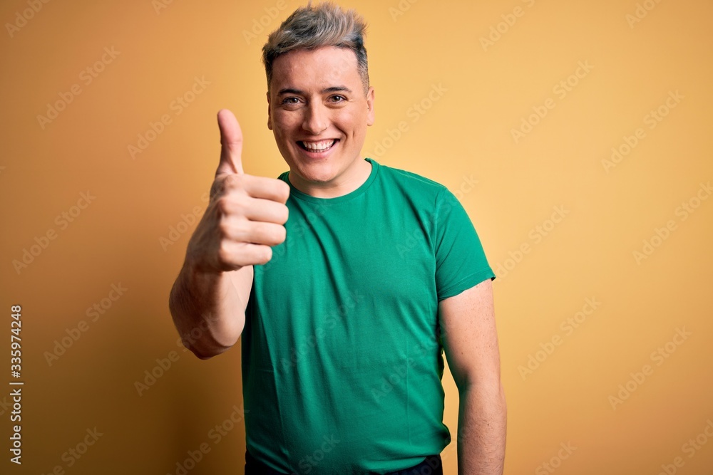 Wall mural young handsome modern man wearing casual green t-shirt over yellow background doing happy thumbs up 