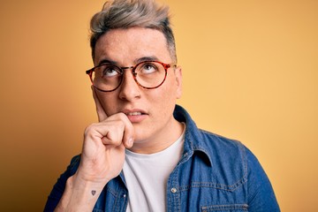 Close up of young handsome modern man wearing glasses and denim jacket over yellow background serious face thinking about question, very confused idea