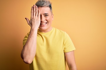 Young handsome modern man wearing yellow shirt over yellow isolated background covering one eye with hand, confident smile on face and surprise emotion.