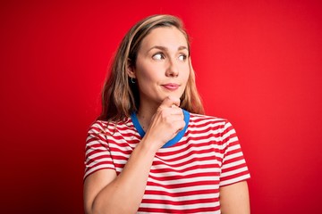 Young beautiful blonde woman wearing casual striped t-shirt over isolated red background with hand on chin thinking about question, pensive expression. Smiling with thoughtful face. Doubt concept.