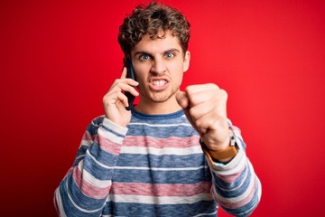 Young blond man with curly hair having conversation talking by the smartphone annoyed and frustrated shouting with anger, crazy and yelling with raised hand, anger concept