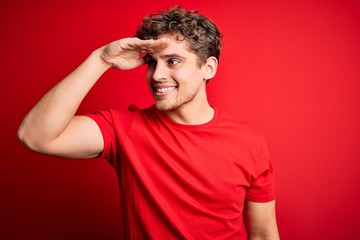 Young blond handsome man with curly hair wearing casual t-shirt over red background very happy and smiling looking far away with hand over head. Searching concept.