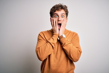 Young blond handsome man with curly hair wearing casual sweater over white background afraid and...