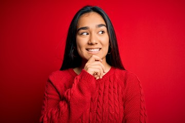 Young beautiful asian woman wearing casual sweater standing over isolated red background with hand on chin thinking about question, pensive expression. Smiling with thoughtful face. Doubt concept.