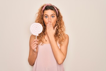 Young beautiful blonde woman with blue eyes eating sweet candy lollipop over pink background cover...