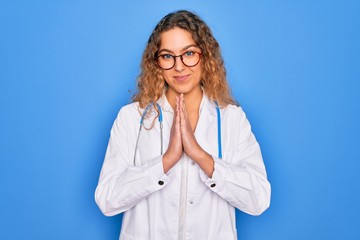 Young beautiful blonde doctor woman with blue eyes wearing coat and stethoscope praying with hands together asking for forgiveness smiling confident.