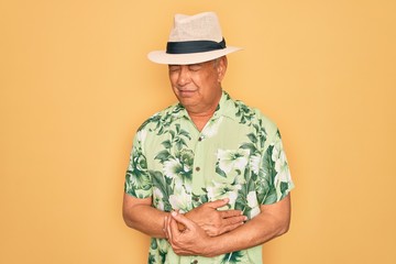 Middle age senior grey-haired man wearing summer hat and floral shirt on beach vacation with hand on stomach because indigestion, painful illness feeling unwell. Ache concept.
