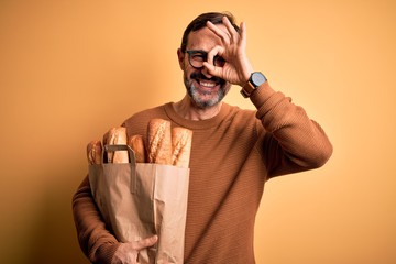 Middle age hoary man holding paper bag with bread standing over isolated yellow background with happy face smiling doing ok sign with hand on eye looking through fingers