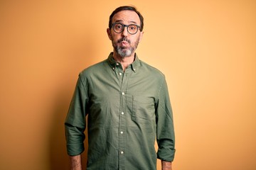 Middle age hoary man wearing casual green shirt and glasses over isolated yellow background puffing cheeks with funny face. Mouth inflated with air, crazy expression.