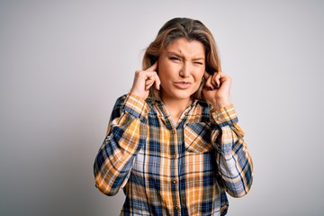 Young beautiful blonde woman wearing casual shirt standing over isolated white background covering ears with fingers with annoyed expression for the noise of loud music. Deaf concept.