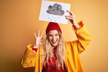 Young beautiful blonde woman wearing raincoat and wool cap holding banner with cloud image doing ok sign with fingers, excellent symbol