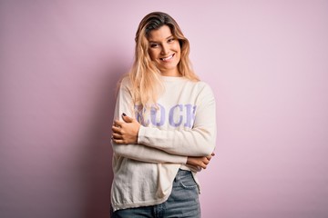 Young beautiful blonde woman wearing casual sweater standing over pink background happy face smiling with crossed arms looking at the camera. Positive person.