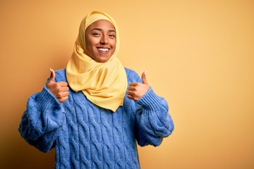 Young African American afro woman wearing muslim hijab over isolated yellow background success sign doing positive gesture with hand, thumbs up smiling and happy. Cheerful expression and winner