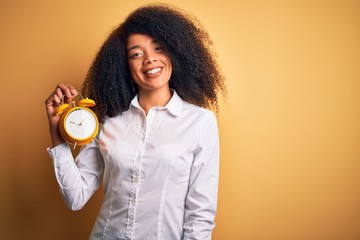 Young african american woman with afro hair holding classic alarm clock over yellow background with a happy face standing and smiling with a confident smile showing teeth