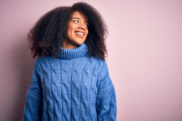 Young beautiful african american woman with afro hair wearing winter sweater over pink background looking away to side with smile on face, natural expression. Laughing confident.