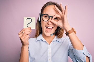 Beautiful call center agent woman working using headset holding reminder with question mark with happy face smiling doing ok sign with hand on eye looking through fingers
