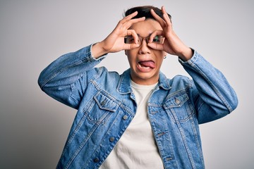 Young handsome chinese man wearing denim jacket and glasses over white background doing ok gesture like binoculars sticking tongue out, eyes looking through fingers. Crazy expression.