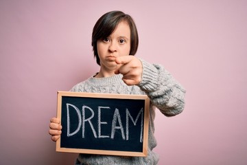 Young down syndrome woman holding blackboard with dream word as message of happiness pointing with finger to the camera and to you, hand sign, positive and confident gesture from the front