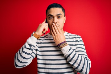 Brazilian man having conversation talking on the smartphone over isolated red background cover mouth with hand shocked with shame for mistake, expression of fear, scared in silence, secret concept