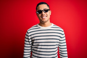 Young brazilian man wearing funny thug life sunglasses over isolated red background with a happy and cool smile on face. Lucky person.