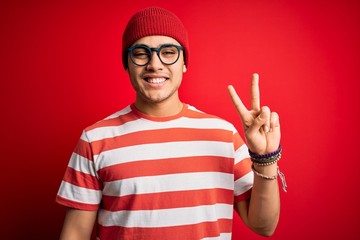 Naklejka premium Young brazilian man wearing casual striped t-shirt and glasses over isolated red background smiling with happy face winking at the camera doing victory sign. Number two.