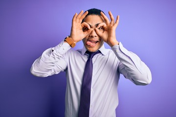 Young brazilian businessman wearing elegant tie standing over isolated purple background doing ok gesture like binoculars sticking tongue out, eyes looking through fingers. Crazy expression.