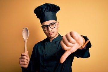 Young brazilian chef man wearing cooker uniform and hat holding wooden spoon with angry face,...