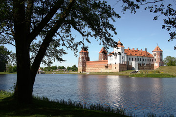 Obraz premium Sunny spring day. In the distance behind the lake ancient Mir Castle Complex is visible. Around fresh greens of leaves and grass.