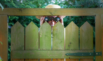 An older man with a straw hat and binoculars looks curiously over a garden fence. Concept:...