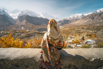A woman wearing traditional dress sitting on wall and looking at Hunza valley in autumn season, Gilgit Baltistan in Pakistan - obrazy, fototapety, plakaty