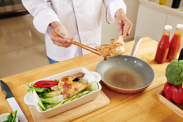 Man using chopsticks and wooden spatula to take fried chicken thighs from frying pan and put it in baking tray with vegetables