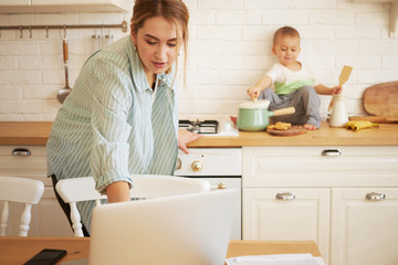 Attractive young mother in casual clothes cooking dinner in kitchen browsing internet on laptop,...