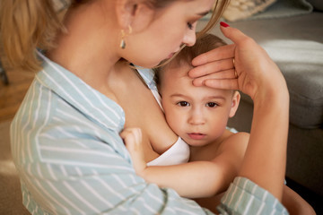 Cropped shot of worried young woman holding her sad infant son, checking if he has fever without...