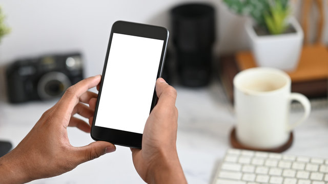 Cropped image of hands holding a white blank screen smartphone over keyboard, coffee cup and office equipment as background. Orderly workspace concept.