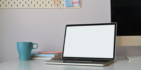 Photo of computer laptop with white blank screen putting on white working desk with coffee cup, stack of book, personal computer and keyboard over orderly living room wall as background.