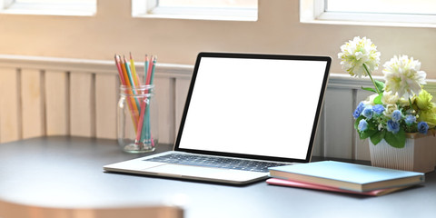 Blank screen computer laptop putting on working desk with bunch of flowers, notebook and pencil holder over comfortable living room as background. Orderly workspace concept.
