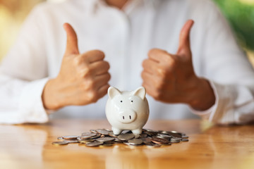 A woman making and showing thumbs up hand sign with coins and piggy bank on the table for saving...