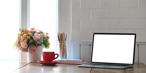Photo of white blank screen computer laptop putting on working desk with bunch of flowers, notebook and pencil holder over comfortable living room as background. Orderly workspace concept.