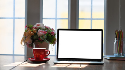 Mockup laptop computer putting on working desk with bunch of flowers, notebook and pencil holder over comfortable wooden table.