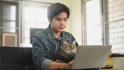 Man carrying an adorable maine coon cat while using a computer laptop and sitting at the leather couch over comfortable living room as background.