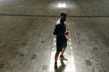 Serious African-American jogger standing in tunnel and drinking vitamin water after running