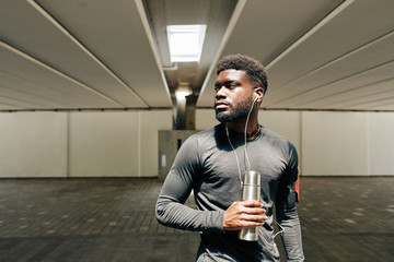Portrait of serious young Black sportsman listening to music with earbuds and drinking water