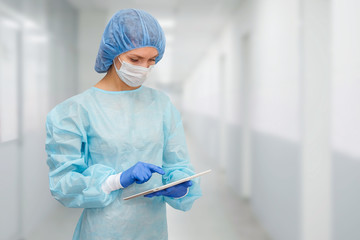 Female doctor in protective suit: medical mask, hat and robe. The woman uses a digital tablet and...