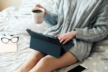 Obraz premium Young woman working on tablet and drinking coffee in bed at home. Cozy, hygge style. Online shopping, working home, freelance, online learning, studying concept. Distance education