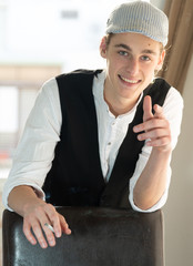 A young man in a cap and vest smokes by the window