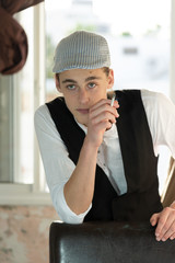 A young man in a cap and vest smokes by the window