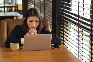  stress young Asian woman in black suit sitting and put her chin on her hands while watching a laptop on wooden table with cellphone and coffee cup