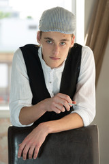 A young man in a cap and vest smokes by the window