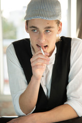 A young man in a cap and vest smokes by the window